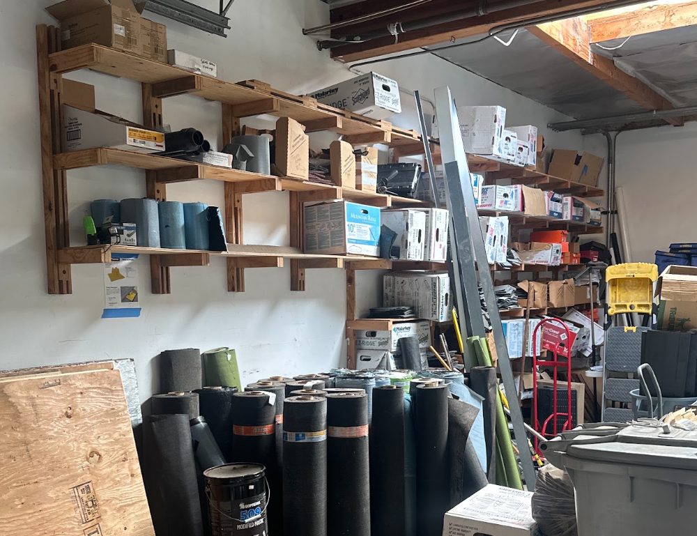 Shelves of roofing supplies and rolls of roofing membrane stacked on the floor inside the McClain Roofing shop.